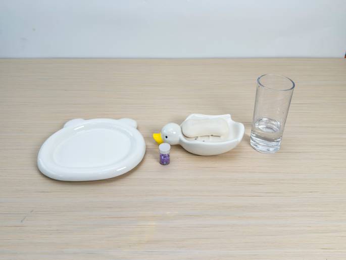 Arranged on a light wood-grain surface are several items, likely for a crafting or experiment setup. A plain white, bear-shaped dish sits to the left. Next to it is a duck-shaped soap dish containing a bar of white soap. A small, clear container filled with purple glitter sits beside the soap dish. A clear glass of water completes the arrangement on the right. The scene suggests preparation for a craft project or a demonstration involving soap, water, and glitter.