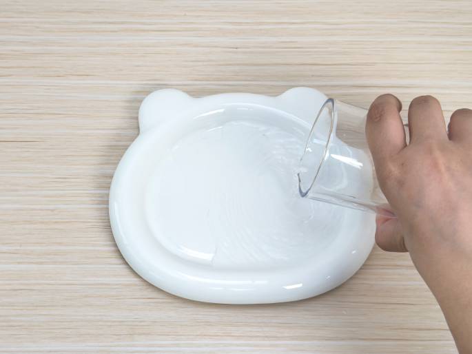 A hand pours water from a clear glass into a white, bear-shaped dish on a light wood-grain table. The water is flowing into the dish, creating ripples. The image likely depicts a step in a science experiment, a crafting activity, or a demonstration involving water.