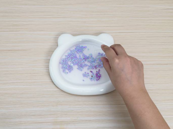 A hand sprinkles purple glitter into a white, bear-shaped dish containing water. The dish rests on a light wood-grain table. The image likely depicts a step in a craft project or experiment involving water and glitter.
