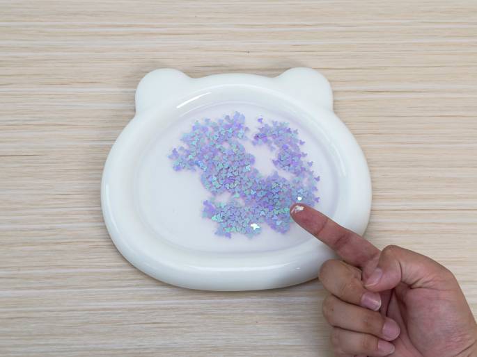 A white, bear-shaped dish filled with water and light purple glitter sits on a light wood-grain table. A finger points towards the glitter in the dish. This image likely illustrates a step in a craft activity or experiment.