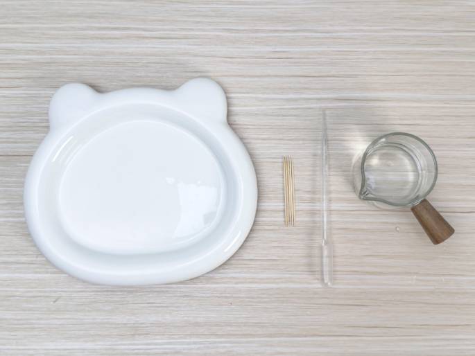 An overhead shot displays several items arranged on a light wood-grain surface. These include a white plate with small ear-like protrusions, a small pile of wooden toothpicks, a clear rectangular glass or acrylic sheet, and a small glass creamer or pitcher with a wooden handle containing clear liquid, likely water. The arrangement suggests preparation for a craft, experiment, or possibly a food preparation scenario.