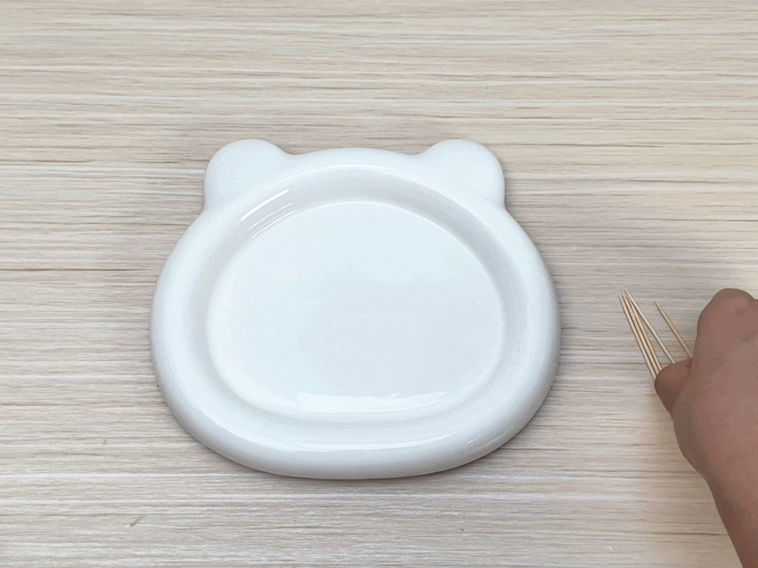 A hand reaches into the frame and places a small group of wooden toothpicks onto a light wood-grain surface. A white plate with small ear-like protrusions is positioned near the toothpicks. The scene suggests the beginning of a craft, experiment, or activity using these simple items.