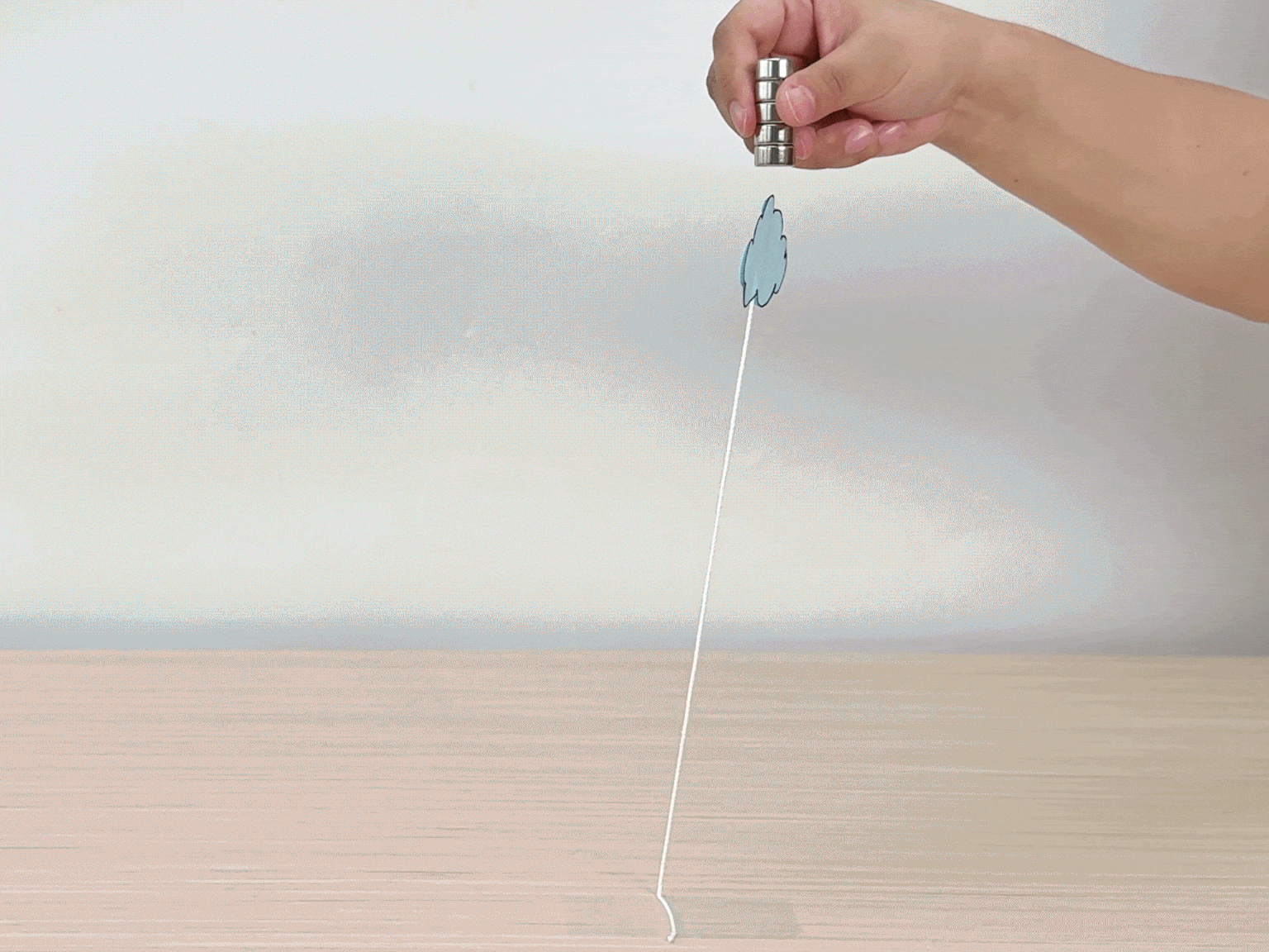 A hand holds a stack of magnets above a paper cloud attached to a string. The cloud is suspended in the air, seemingly floating, due to the magnetic force repelling it from the magnets held above. This demonstrates a simple physics experiment involving magnetism.