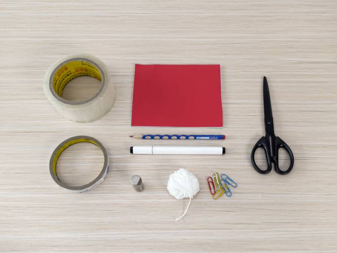 An assortment of craft supplies lies on a wooden table. These include rolls of clear and brown tape, a red piece of paper or cardstock, a pencil, a white marker, scissors, a small magnet, some white yarn or string, and several colorful paper clips. These items suggest preparation for a craft project.