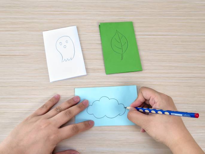 On a light wood-grain surface, two hands are engaged in a crafting activity. In the background, a white piece of paper features a simple drawing of a ghost, and a green piece of paper displays a drawing of a leaf. In the foreground, a hand holds a light blue piece of paper while the other hand, holding a blue and white polka-dotted pencil, draws a cloud shape on it. The scene suggests a paper crafting or drawing project.