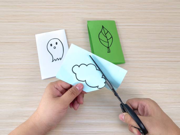 Two hands work on a crafting project on a light wood-grain table. One hand holds a light blue piece of paper with a drawn cloud, while the other hand uses scissors to cut out the cloud shape. In the background, there's a white paper with a ghost drawing and a green paper with a leaf drawing, suggesting a themed crafting activity.
