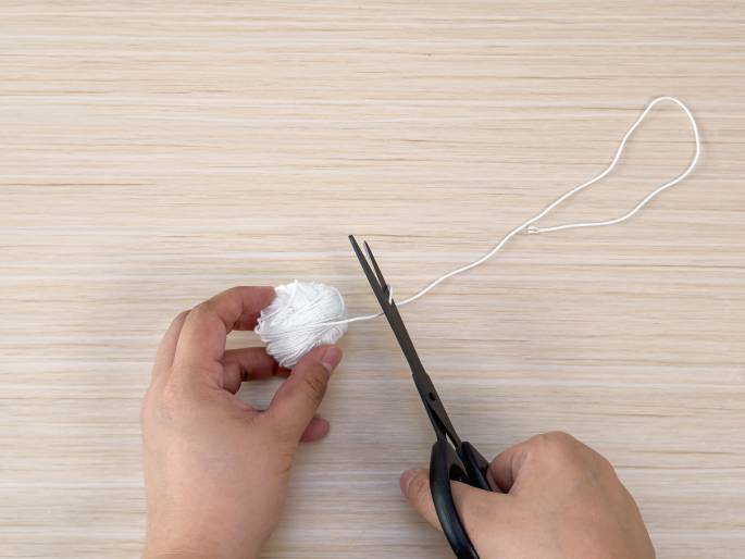 Two hands are shown on a light wood-grain surface working with a small ball of white yarn and a pair of black scissors. One hand holds the yarn, from which a length of string extends. The other hand holds the scissors and is in the process of cutting the string. This image likely depicts a step in a crafting or DIY project.