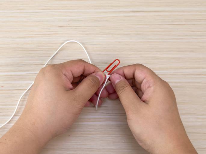 Two hands manipulate a piece of white string on a light wood-grain table. A red paperclip is attached to the string, and the hands appear to be in the process of tying a knot or securing the string with the paperclip. This suggests a step in a crafting, DIY, or household task.