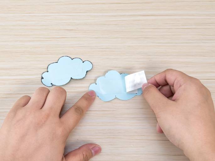 On a light wood-grain surface, two hands interact with light blue cloud-shaped cutouts. One hand points towards a completed cloud, while the other hand peels the backing off of an adhesive strip attached to another cloud cutout. This suggests a crafting or decorating activity involving adhesive stickers.
