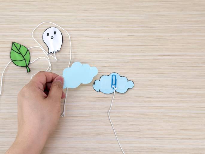 A hand interacts with a crafting project on a light wood-grain table. Several small paper cutouts, including a ghost, a leaf, and two light blue clouds, are attached to lengths of white string. The hand holds one of the cloud cutouts. A small blue paperclip is attached to another cloud, suggesting they are being linked or hung together. The scene depicts a simple mobile or hanging decoration being assembled.