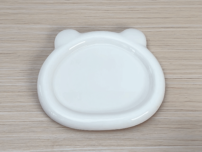 A small, white, bear-shaped dish sits on a light wooden table. A hand places a small piece of white paper towel with a yellow, powdery substance on it into the dish. A small amount of red powder is already in the bottom of the dish. The dish is then filled with a clear liquid, likely water. The yellow powder dissolves and disperses throughout the liquid, turning it yellow. The red powder also disperses, creating a subtle orange hue in parts of the yellow liquid. This video appears to demonstrate a simple experiment involving solubility and color mixing.