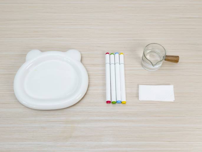 Laid out on a light wood-grain surface are several items: a white plate with small ear-like protrusions, four markers with red, green, blue, and yellow caps, a small clear glass pitcher or creamer with a wooden handle containing clear liquid (likely water), and a folded piece of white paper or cloth. This arrangement suggests preparation for an art project or experiment.