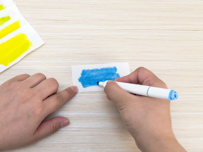 On a light wooden surface, a hand holding a blue marker colors a small piece of white paper or cloth. Another hand's finger points towards the paper, possibly guiding the coloring or indicating a specific area. In the top left corner, another piece of paper is visible with yellow and light green marker streaks, suggesting previous color tests or parts of a larger art activity.