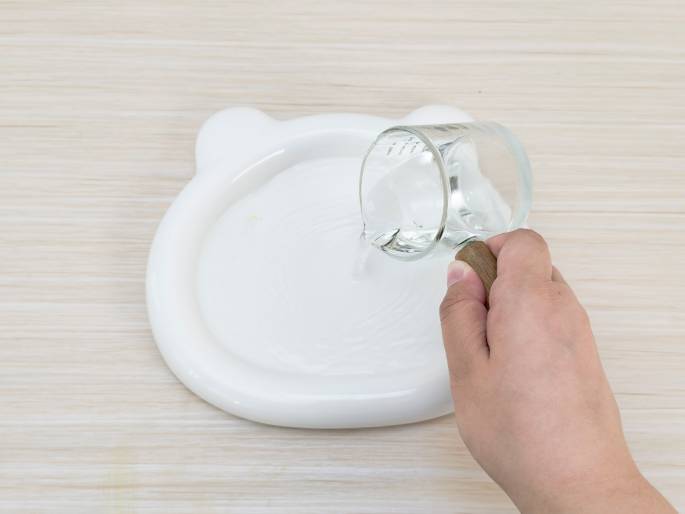 A hand pours clear liquid, presumably water, from a small glass pitcher with a wooden handle onto a white plate with small ear-like protrusions. The plate sits on a light wood-grain surface. The image suggests an experiment, art project, or other activity involving water and the plate.