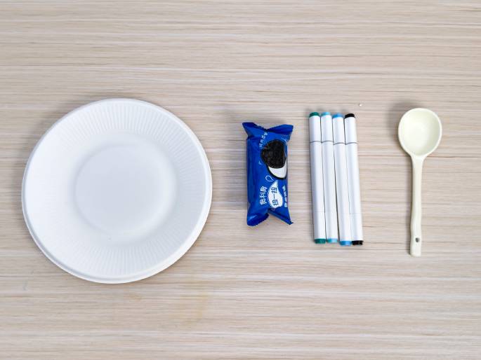 Arranged on a light wood-grain surface are several items, suggesting preparation for a craft or activity. A white paper plate sits to the left. Next to it is a blue-wrapped package, possibly containing a snack or art supply. A group of colorful markers with white bodies lies beside the package, and a white plastic spoon is positioned to the far right. The image conveys a sense of getting ready for a creative project.