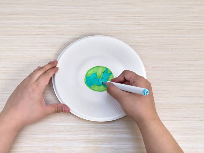 Two hands are engaged in an art project on a light wood-grain table. One hand steadies a white paper plate, while the other hand uses a light blue marker to color a drawing of the Earth on the plate. The Earth is depicted with green continents and blue oceans. This scene represents a child's art activity or a craft project related to Earth Day or environmental themes.