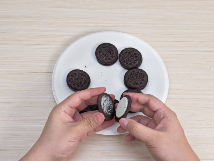 Two hands hold a split Oreo cookie over a white paper plate on a light wood-grain table. Several other whole Oreo cookies are arranged on the plate. One half of the split cookie displays the white cream filling, while the other half has had the filling removed, revealing the dark chocolate cookie surface. This image suggests a common practice of separating Oreo cookies to eat the filling or to use the cookie parts for other purposes, such as crafts or desserts.