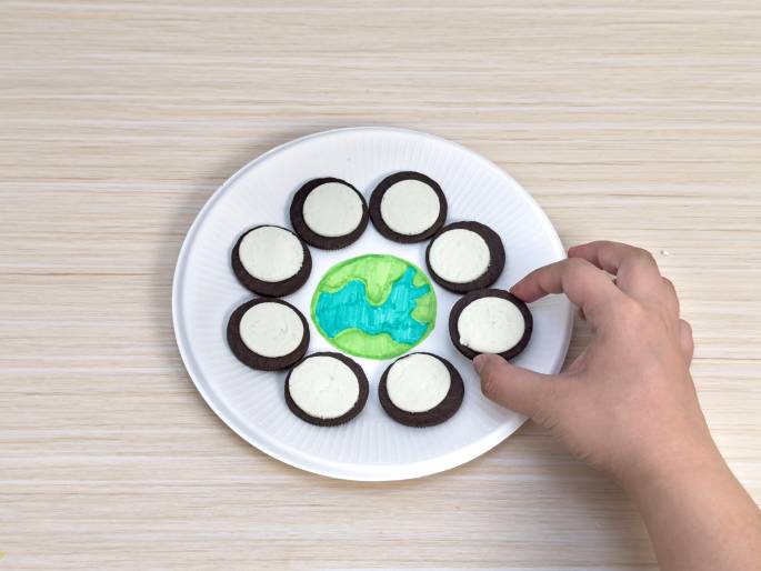 On a light wood-grain table, a white paper plate displays a creative arrangement. A drawing of the Earth, with green continents and blue oceans, is at the center. Around the Earth, several Oreo cookies are placed with their cream filling facing outwards, forming a ring. A hand reaches in to pick up one of the Oreo cookies. This image likely depicts a child's craft project or a fun snack arrangement related to Earth Day or environmental themes.