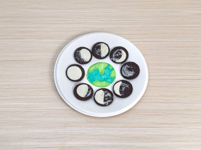 A white paper plate rests on a light wood-grain surface. In the center of the plate, there's a drawing of the Earth with green continents and blue oceans. Arranged around the Earth drawing are Oreo cookies, each with a portion of the white cream filling scraped away to represent the phases of the moon. This display illustrates the different lunar phases using Oreo cookies as a visual aid, likely part of a science project or educational activity.
