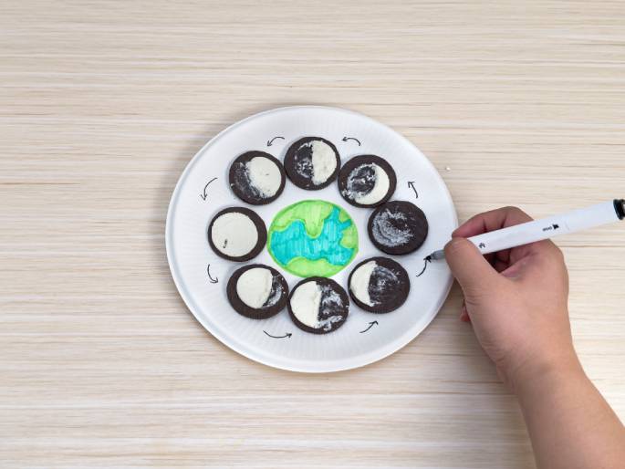 On a light wood-grain table, a white paper plate presents a visual representation of the lunar cycle. A drawing of Earth, depicted with green continents and blue oceans, sits in the center. Oreo cookies encircle the Earth, each with its cream filling partially scraped off to illustrate the different phases of the moon. A hand holding a white marker is drawing arrows around the cookies, indicating the direction of the moon's orbit and the progression through the phases. This setup clearly demonstrates the lunar cycle using Oreo cookies as a learning tool, likely for a science project or educational activity.