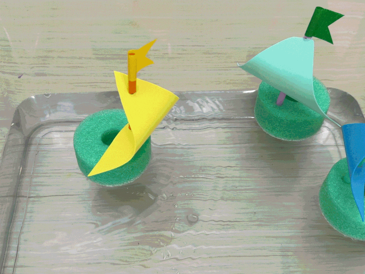 Three small toy sailboats float in a clear container of water. The boats are crafted from green foam rings with colorful paper sails attached to straws. This playful scene evokes a sense of childhood crafts and simple fun.