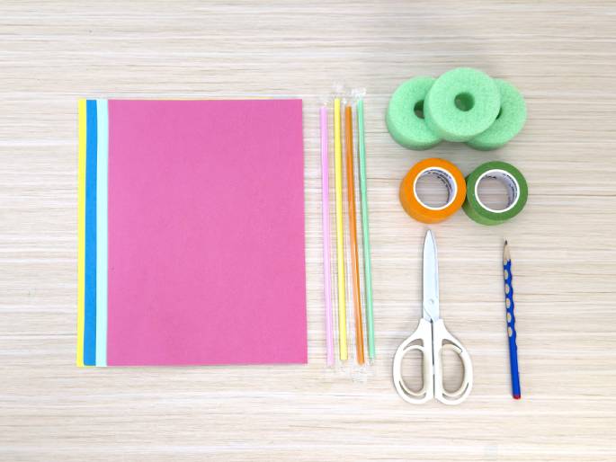An assortment of craft supplies is laid out on a light wood-grain table. The items include a sheet of pink construction paper, several colorful straws, a pair of light green foam rings, rolls of orange and green tape, a pair of white scissors, and a blue pencil. This flat lay arrangement suggests preparation for a children's craft project or DIY activity.