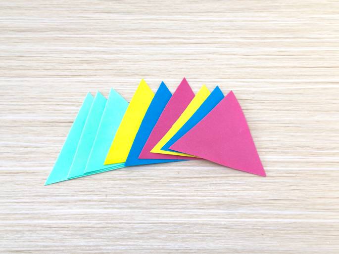 Several colorful paper triangles are arranged on a light wood-grain surface. The triangles fan out from a central point, overlapping slightly. They appear to be cut from construction paper in shades of light teal, bright yellow, blue, and pink. These vibrant triangles likely form part of a craft project or decorative element.