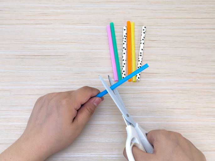 Two hands are shown on a light wood-grain surface. One hand holds a blue plastic drinking straw, while the other hand holds a pair of white scissors and is in the process of cutting the straw. A few other colorful straws, including pink, green, orange, black-and-white patterned, and another blue one, lie on the table near the cutting action. This image likely depicts a step in a craft project or activity involving cutting straws.