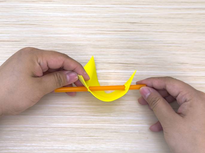 Two hands manipulate a piece of bright yellow construction paper and an orange straw on a light wood-grain table. The paper is folded over the straw, and the hands are pinching the edges of the paper around the straw. This image appears to capture a step in a craft project, likely creating a three-dimensional shape or structure using paper and straws.