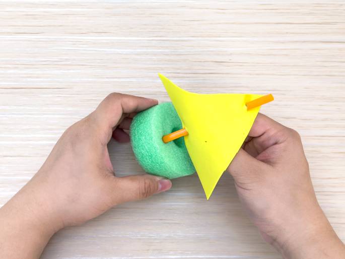 Two hands hold a small, simple sailboat craft on a light wood-grain table. The boat's hull is a light green foam ring, and a short piece of orange straw is inserted through the center. A bright yellow triangle of construction paper, acting as the sail, is attached to the straw. The hands are positioning or adjusting the sail. This image showcases a completed or near-completed children's craft project.