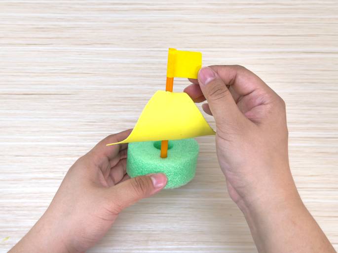 Two hands hold a finished toy sailboat craft on a light wood-grain background. The boat's hull is a light green foam ring. An orange straw mast rises from the center, holding a bright yellow triangular sail. A small, rectangular yellow flag is attached to the top of the mast. One hand holds the base of the boat, while the other gently adjusts the flag. This image showcases a completed children's craft project.