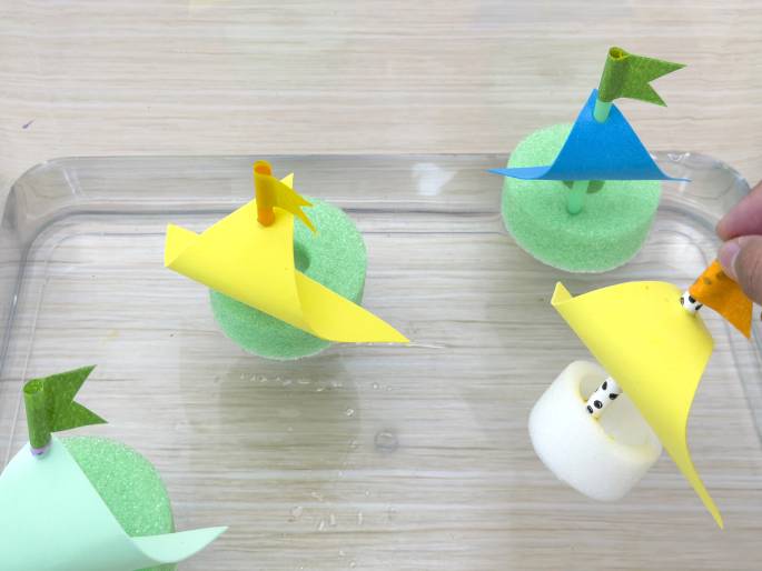 Four small, handmade sailboats float in a clear, shallow rectangular container with a little water. Each boat uses a light green foam ring as a hull and has a colored paper sail attached to a straw mast with a small paper flag at the top. The sails and flags vary in color: light green with a dark green flag, yellow with an orange flag, blue with a light green flag, and yellow with an orange flag. The last boat has a white foam hull and a black-and-white spotted straw mast. A hand is visible in the top right corner, placing the last boat into the water. This image captures a playful scene of toy sailboats ready to race or sail.