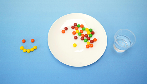 On a pale blue surface, a white plate holds a scattering of multicoloured Skittles sweets. A small smiley face, also made from Skittles, is positioned to the left of the plate. Beside the plate sits a clear plastic cup containing water. The arrangement suggests the beginning of a Skittles science experiment, using the sweets and water to demonstrate colour separation or diffusion. The overall style is simple and clean, highlighting the colourful sweets against the plain background.