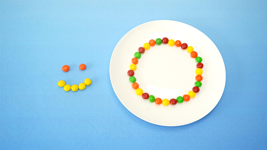 Against a light blue background, a white plate displays a circle of colourful Skittles sweets arranged around its edge. A small smiley face, also constructed from Skittles, is positioned to the plate's left. The colours are bright and distinct, and the arrangement is neat and precise. The image clearly shows the set-up for a Skittles science experiment, likely demonstrating colour separation or diffusion in water. The overall aesthetic is simple, clean, and playful, highlighting the colourful candies.