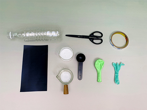 The image shows a collection of items laid out on a light grey surface, clearly prepared for a science experiment, likely involving inflating a balloon using a chemical reaction. The items include a plastic bottle, scissors, adhesive tape, a small black measuring spoon, two small containers of white powder (presumably bicarbonate of soda), a small glass jug containing a clear liquid (likely vinegar), a light green balloon, and a piece of light green pipe cleaner. The arrangement is neat and suggests a planned, step-by-step process. A piece of dark material, possibly card or a similar material, is also included, possibly for creating a funnel or covering the bottle neck.