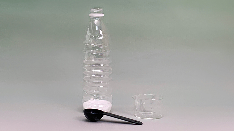 A clear plastic bottle sits on a pale surface against a muted green background. At the base of the bottle is a quantity of white powder, likely bicarbonate of soda. Beside it is a small, clear measuring jug and a black measuring spoon. The scene is simply lit and the objects are arranged in a straightforward manner, suggesting the preparation for a science experiment, possibly involving the inflation of a balloon using a chemical reaction.