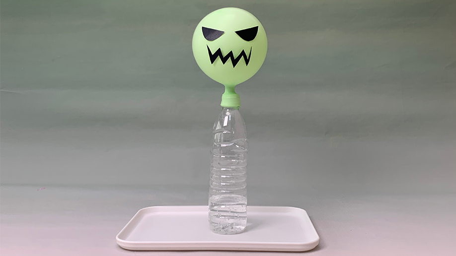 A light green balloon, decorated with a simple black face to resemble a ghost, sits atop a clear plastic bottle partially filled with water. The bottle is placed on a white tray. The background is a soft, muted green. This setup suggests a science experiment, possibly demonstrating the production of carbon dioxide gas to inflate the balloon, creating a fun and spooky Halloween-themed activity.