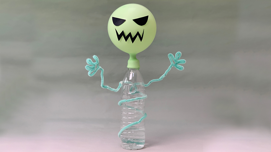 A playful, ghostly figure is constructed from a light green balloon with a drawn-on face, attached to a clear plastic bottle partially filled with water. Two light teal pipe cleaners are shaped and attached to form arms, giving the creation a whimsical, spooky appearance. The bottle is wrapped with a further length of pipe cleaner, adding to the overall effect. The background is a soft, muted green, complementing the colours of the ghost figure. The image suggests a fun, creative science project, possibly involving a chemical reaction to inflate the balloon.