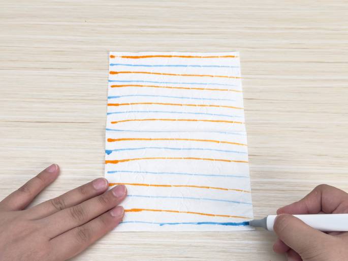 On a light wood-grain surface, a hand draws horizontal blue lines with a marker on a piece of white paper or cloth. The paper already has alternating orange and light blue horizontal lines drawn on it, folded slightly horizontally near the center. Another hand is positioned on the left side of the paper, appearing to hold it in place. The scene suggests an art activity or experiment involving color and lines.