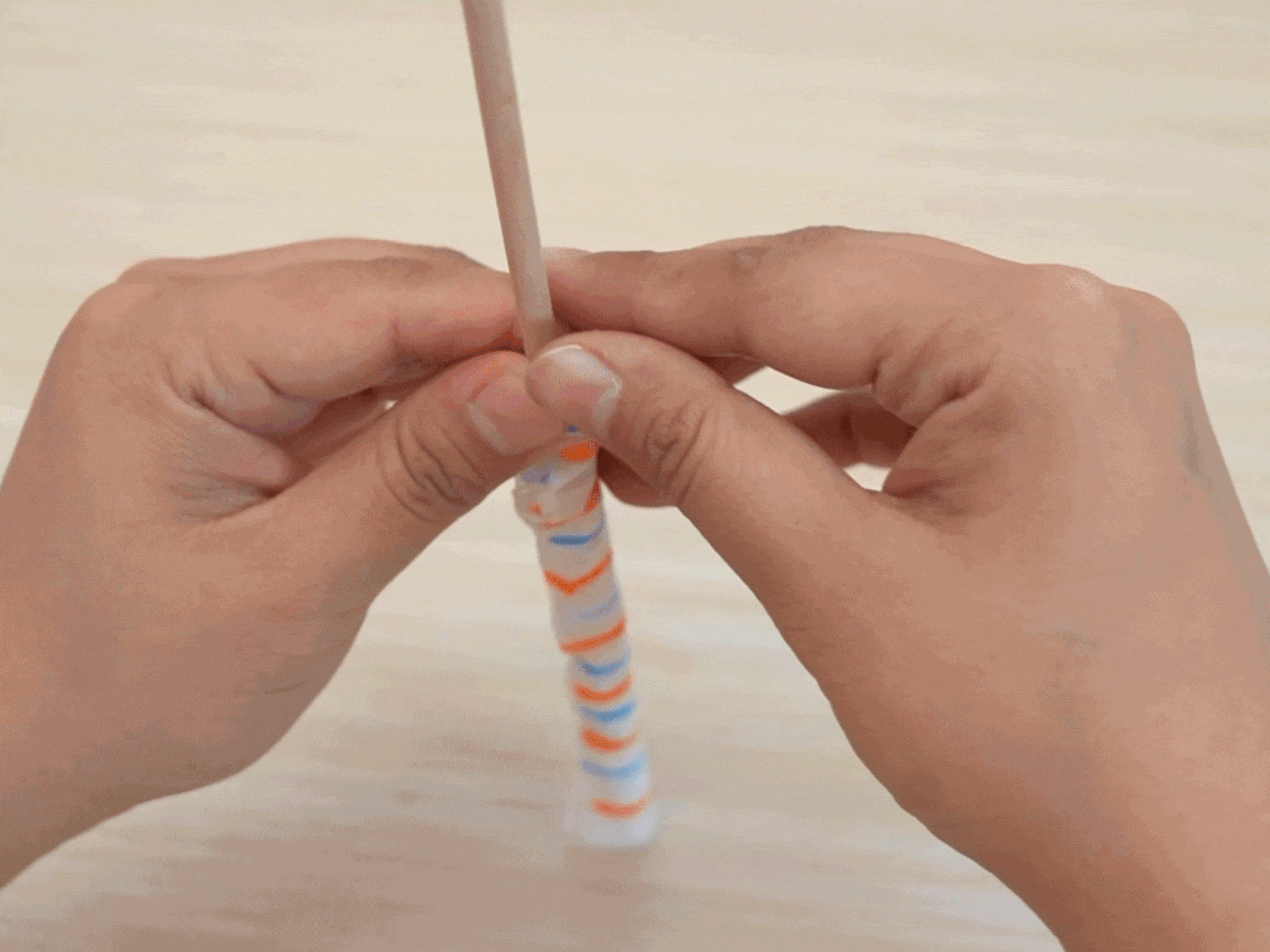 Two hands carefully roll a piece of white material with orange and blue stripes around a thin wooden dowel. The material appears to be paper or cloth, possibly glued, as it adheres to the dowel and itself as it is rolled. This suggests the creation of a small craft or decorative item.