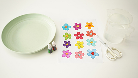 A pale green shallow dish, two small bottles of coloured liquid, a sheet of paper with drawn flowers, a measuring jug of water, and a pair of scissors are arranged on a white surface.