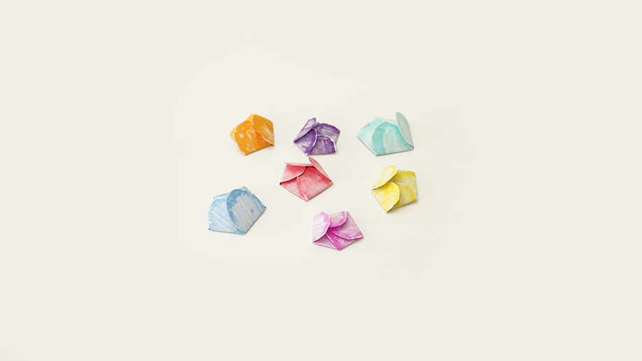 Several small, folded paper shapes in assorted pastel colours are scattered on a white surface.