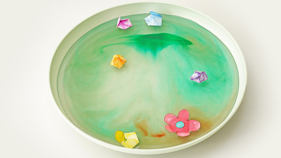 A pale green shallow dish contains green-tinged water in which several small, folded paper shapes and one unfolded paper flower float.
