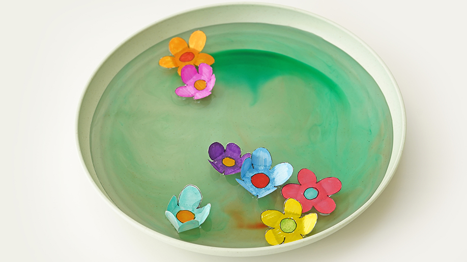 Several colourful paper flowers float on the surface of green-tinged water in a pale green shallow dish.