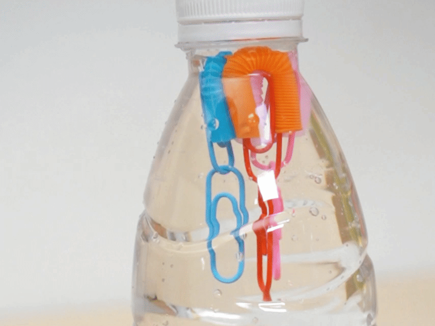 A clear plastic bottle is partially filled with water. Colorful paper clips are linked together and inserted into the bottle's opening, dangling down into the water. The setup suggests a simple science experiment or demonstration, possibly related to buoyancy or water displacement.