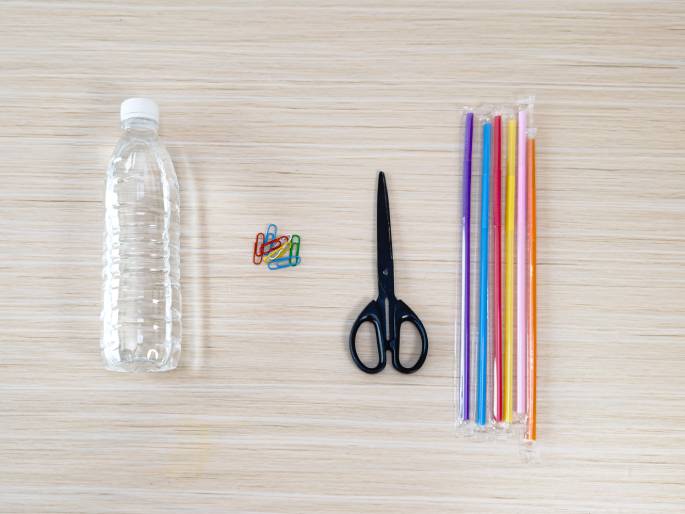 A flat lay arrangement of craft supplies on a light wood-grain table. A clear plastic water bottle lies on its side on the left. A small pile of colorful paper clips sits in the center. A pair of black scissors is positioned to the right of the paper clips. A package of colorful plastic straws, still in its plastic wrap, is on the far right. These items suggest preparation for a craft project involving these materials.
