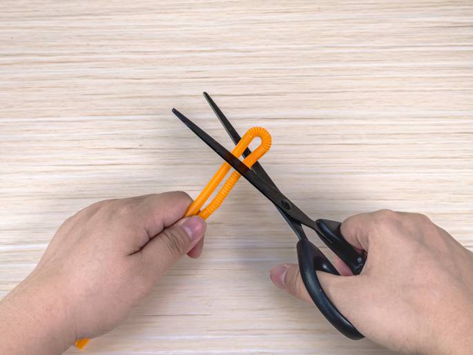 Two hands are shown on a light wood-grain background. One hand holds an orange flexible drinking straw that has been bent into a small loop at the top. The other hand holds a pair of black scissors, positioned to cut just below the looped portion of the straw. The image clearly depicts the process of cutting a drinking straw with scissors.