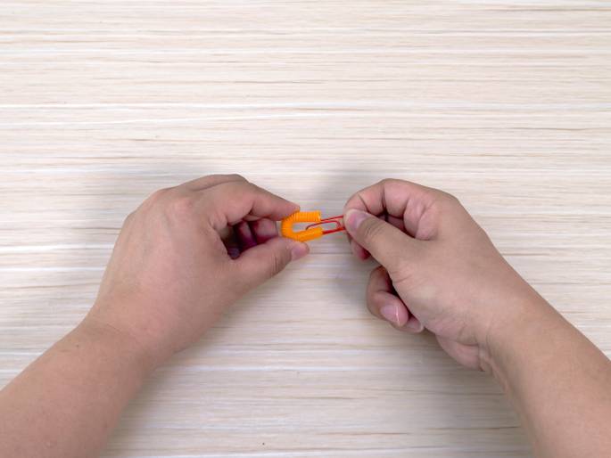 Two hands manipulate a small orange section of a flexible drinking straw and a red paperclip against a light wood-grain background. The straw piece, bent into a loop, has the paperclip clipped onto it. The hands appear to be in the process of attaching or securing these two small items together. This close-up image likely demonstrates a step in a craft project.
