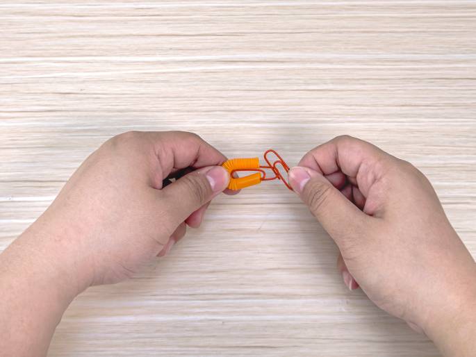 Two hands hold a small, looped piece of orange flexible straw with a red paperclip attached to it against a light wood-grained background. The paperclip is partially open, and the hands seem to be adjusting or demonstrating how the paperclip interacts with the straw loop. This image likely illustrates a step in a simple craft or experiment.
