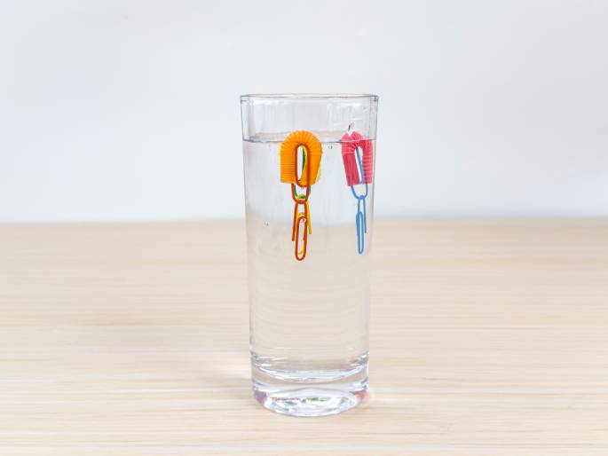 A clear glass of water sits on a light wood-grain table against a plain white background. Two small, looped pieces of flexible drinking straw, one orange and one pink, float on the surface of the water. Each straw piece has a paperclip – one multicolored and one light blue – attached. This image likely depicts a simple science experiment demonstrating buoyancy or surface tension.